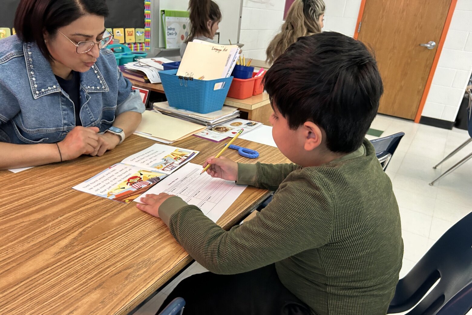 student works at table with teacher across